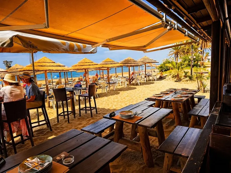 Wooden tables under the awning, with thatched parasols and the sea beyond