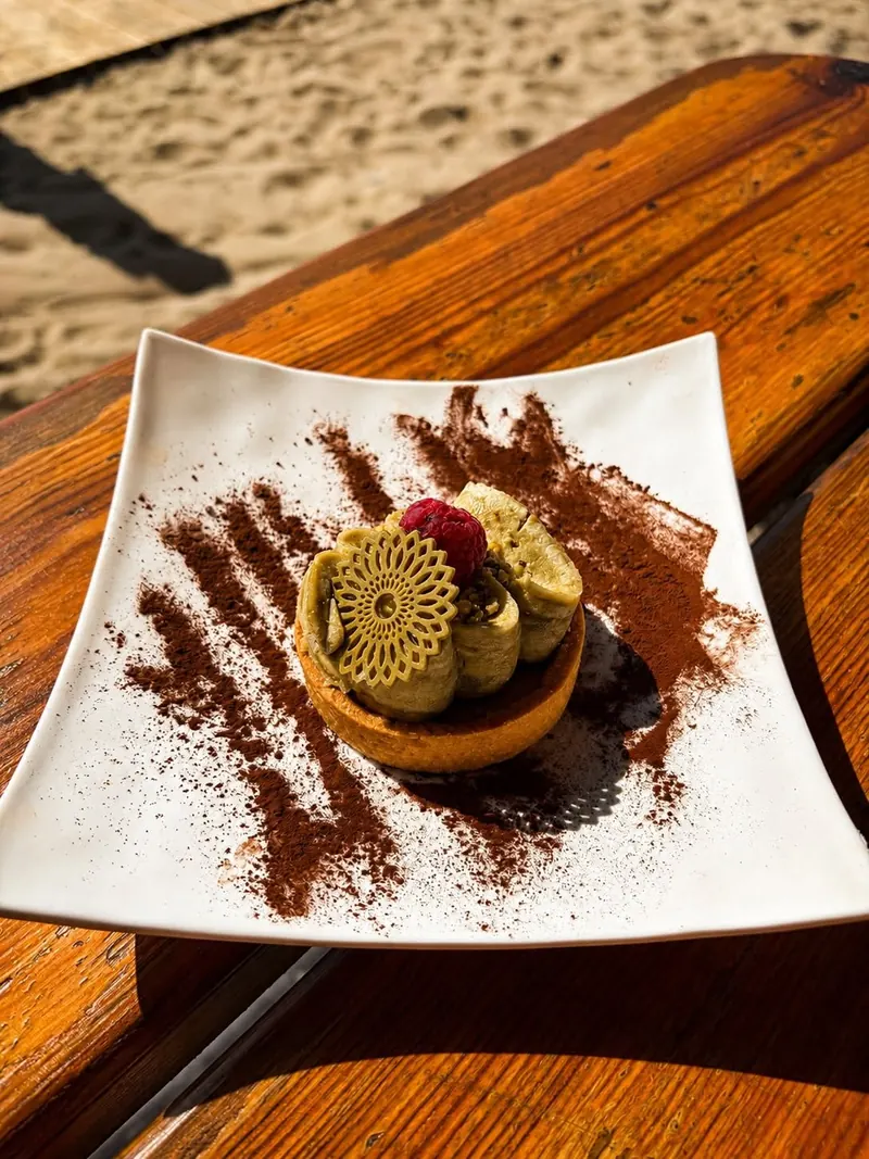 A tartlet with a green pistachio-style topping and a raspberry, on a sunlit wooden table