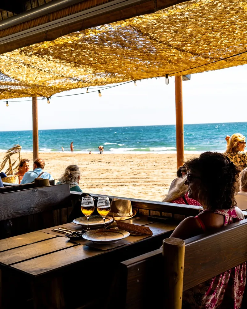 Guests at wooden tables under the reed shade, looking out over the beach and the sea