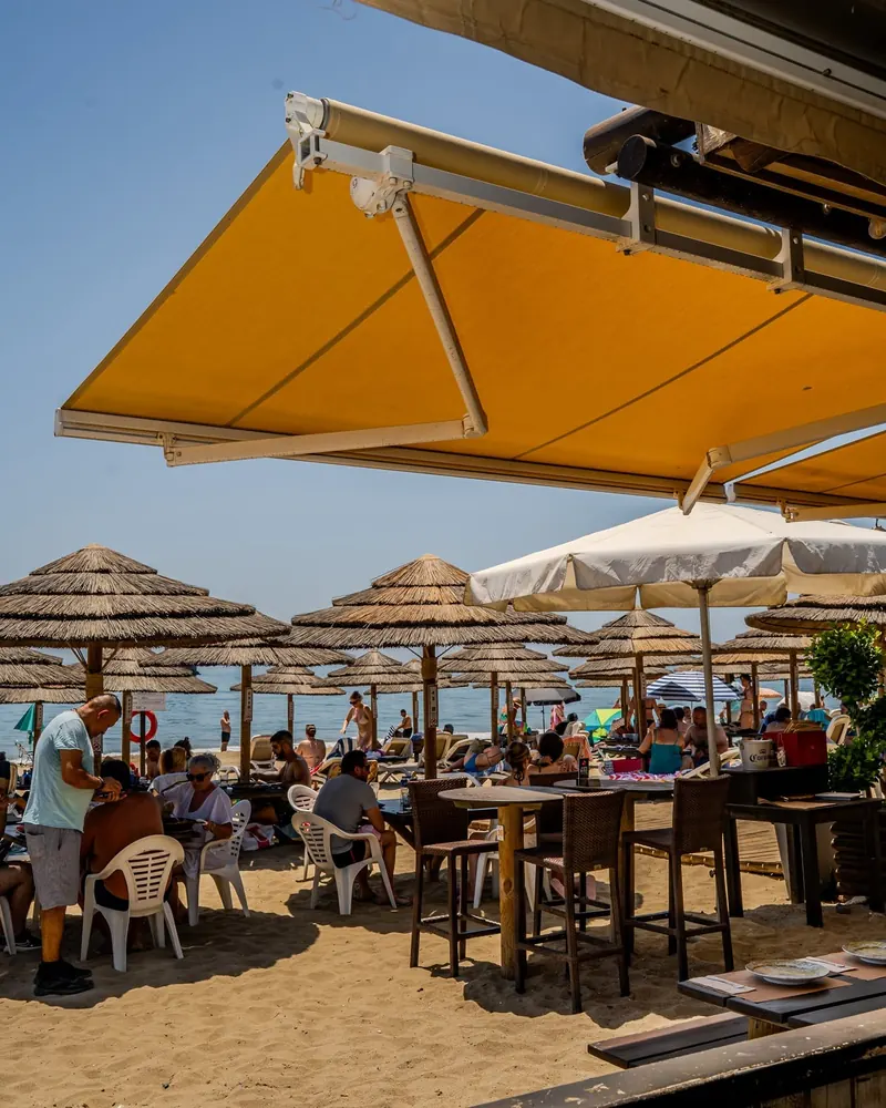 Terrace tables under the yellow awning, thatched parasols across the sand